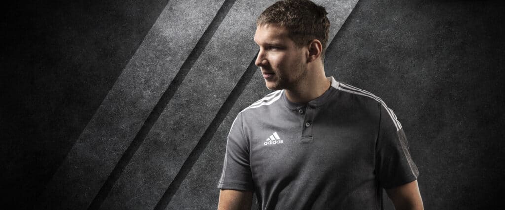 Sportler posiert in grauem adidas Poloshirt Tiro 21 vor grauem Betonhintergrund.